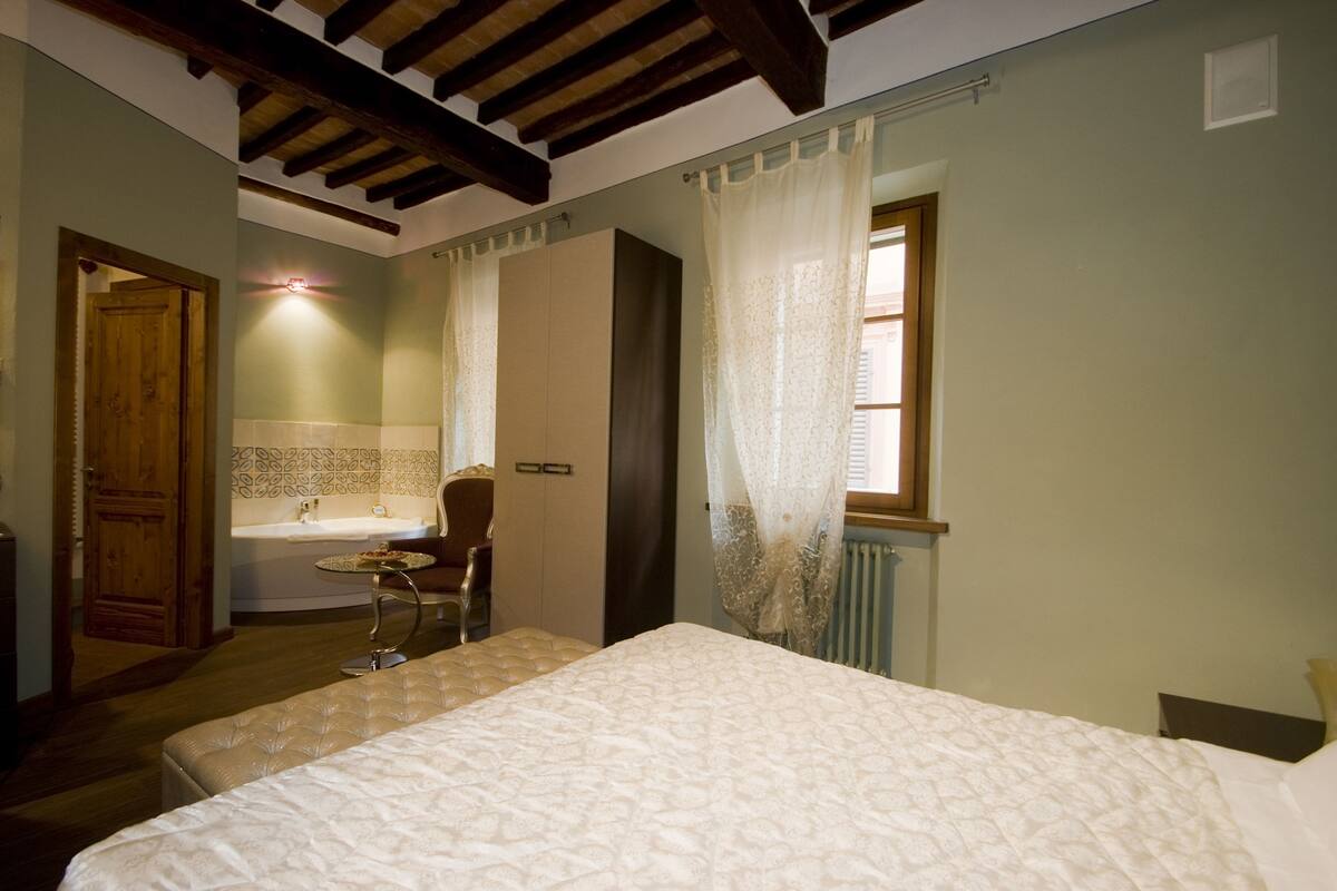 Une chambre à coucher comprenant un lit, une chaise, une petite table, une porte en bois, une fenêtre avec des rideaux, et une salle de bain avec un lavabo et un miroir.
