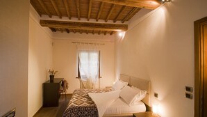 Une chambre avec un plafond en bois, un lit recouvert de draps blancs, une table de chevet avec une lampe et une fenêtre ornée de rideaux légers.