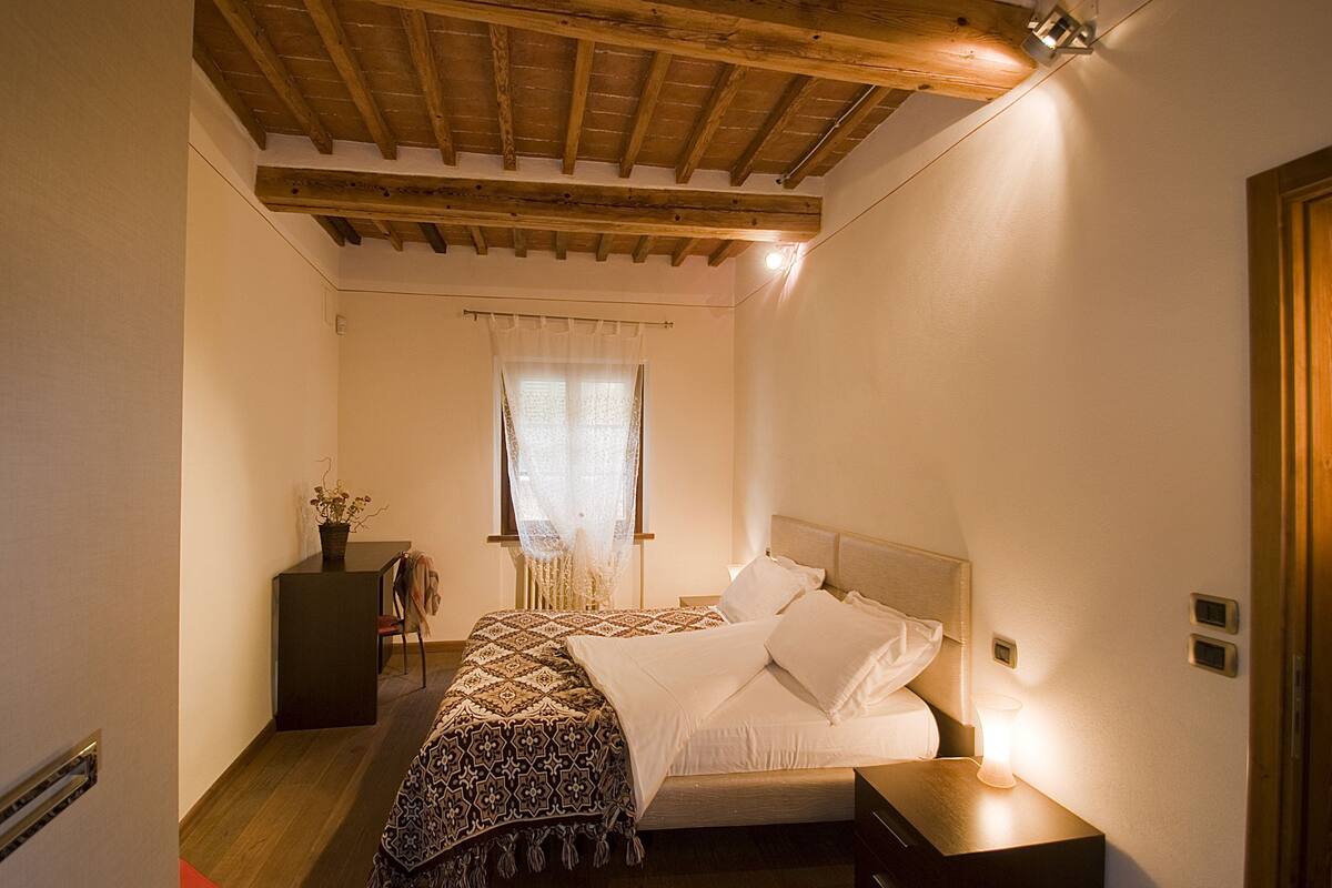 Une chambre avec un plafond en bois, un lit recouvert de draps blancs, une table de chevet avec une lampe et une fenêtre ornée de rideaux légers.