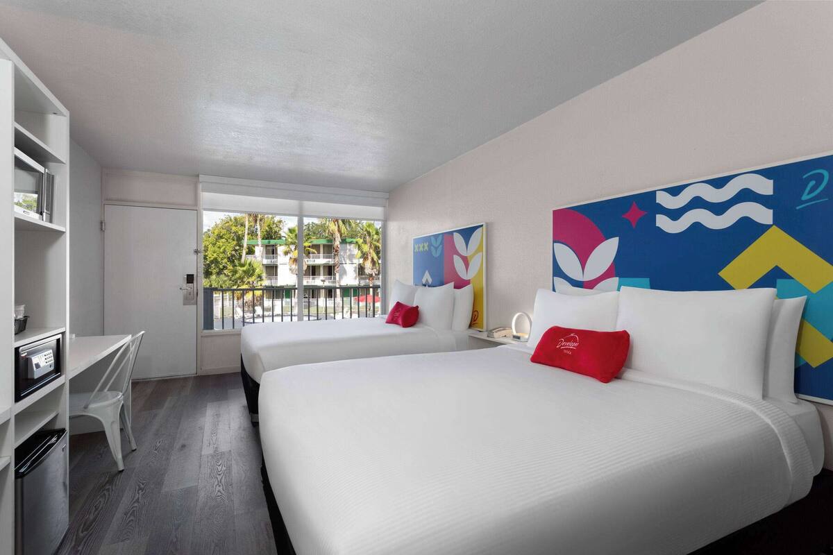 A hotel room with two beds, a desk, and a balcony with a view of buildings and trees.