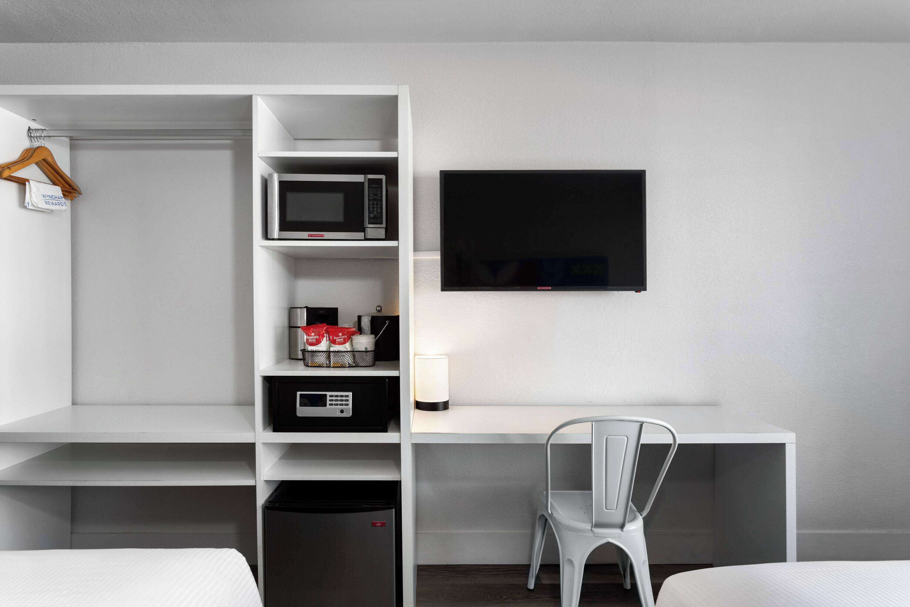A modern hotel room with a white desk, a flat-screen TV, a microwave, a mini-fridge, and a chair.