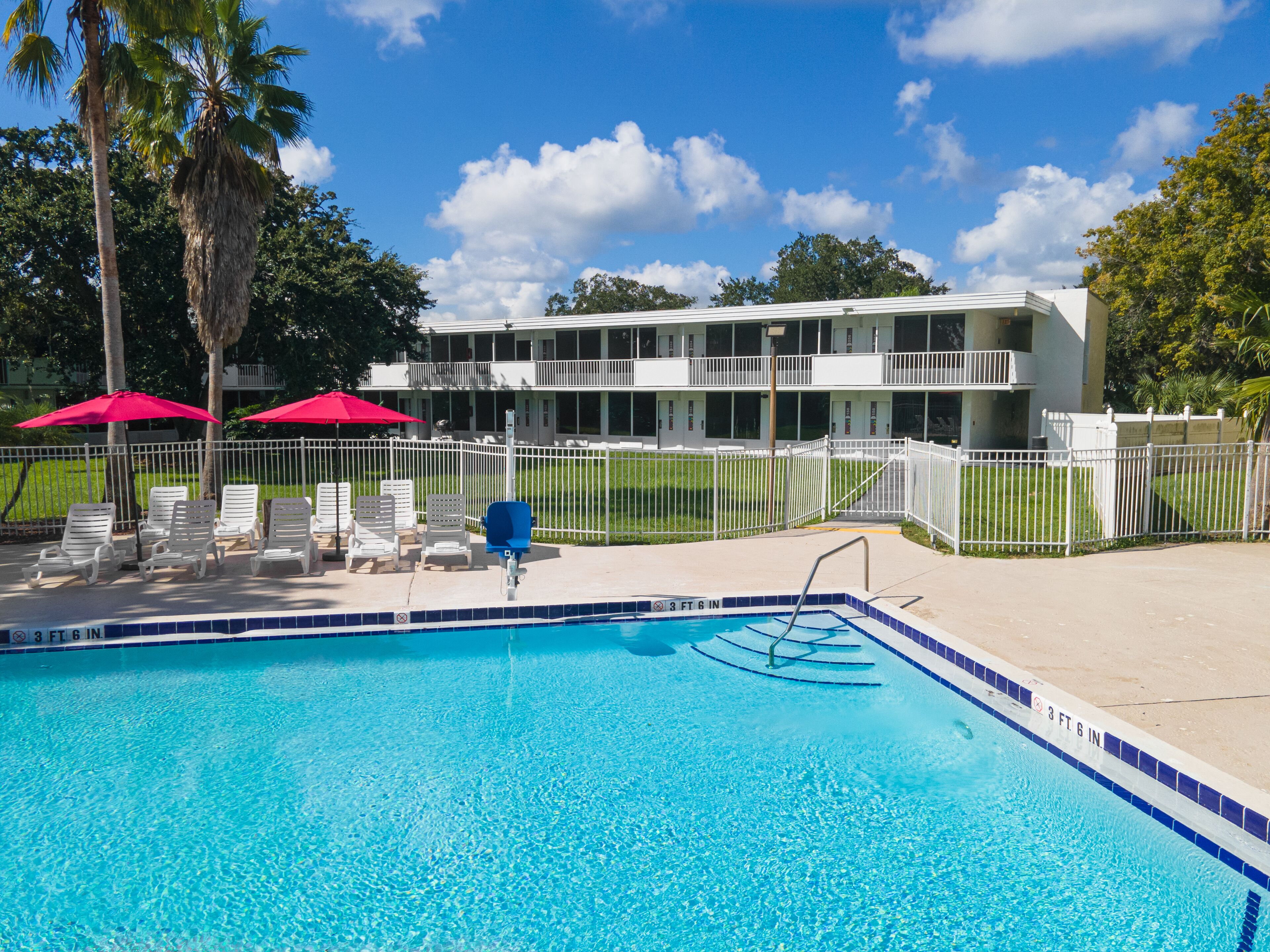 A swimming pool with lounge chairs and umbrellas, surrounded by a white fence and a building with balconies.