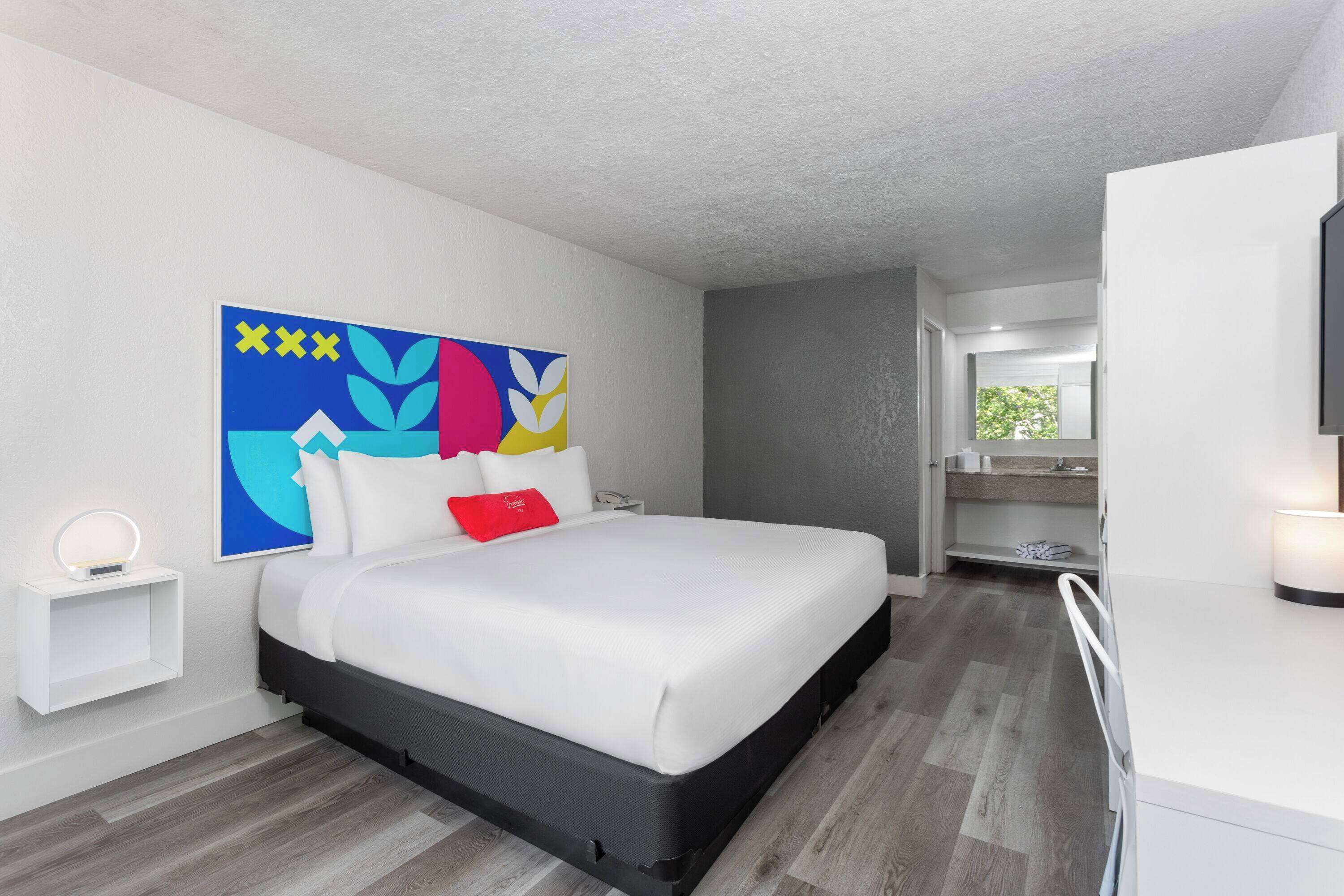 A modern hotel room with a bed, a bedside table with a lamp, a large abstract painting, and a view of the bathroom.