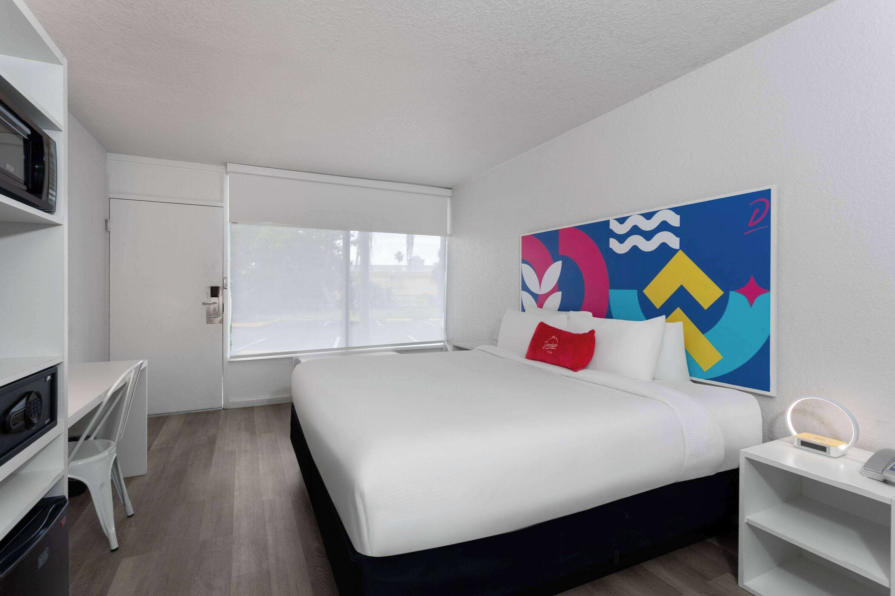 Una habitació d'hotel moderna amb un llit, una taula de treball, una cadira, un televisor i una finestra amb persianes.