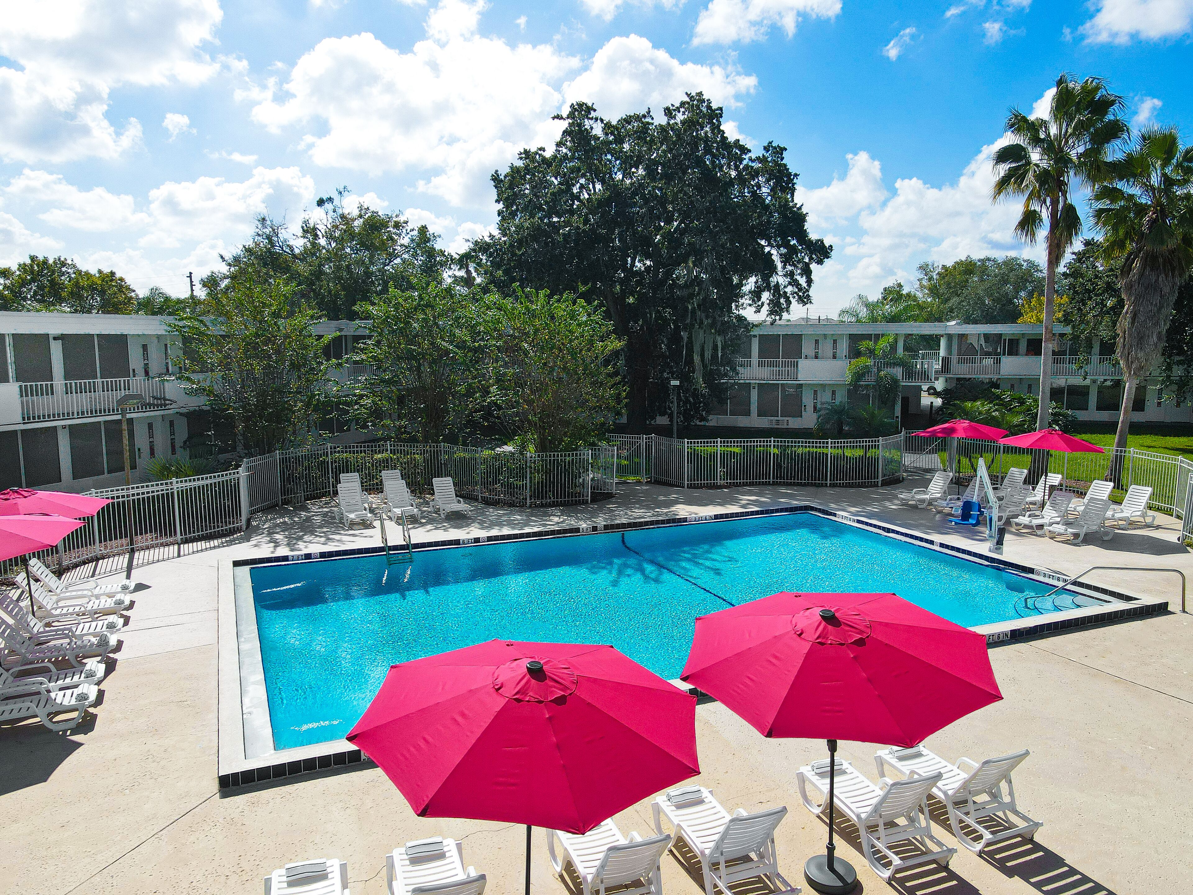 A swimming pool with sun loungers and pink umbrellas, surrounded by a fence and trees.