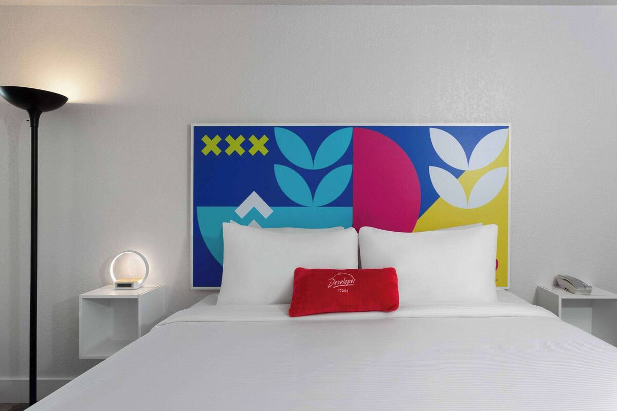 A hotel room with a bed, a bedside lamp, a nightstand, a phone, and a colorful abstract artwork on the wall.