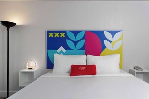 A hotel room with a bed, a bedside lamp, a nightstand, a phone, and a colorful abstract artwork on the wall.