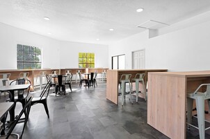 A modern, well-lit room with white walls, grey tiled flooring, and a mix of black and white chairs. There are tables and a wooden counter, with large windows providing natural light.