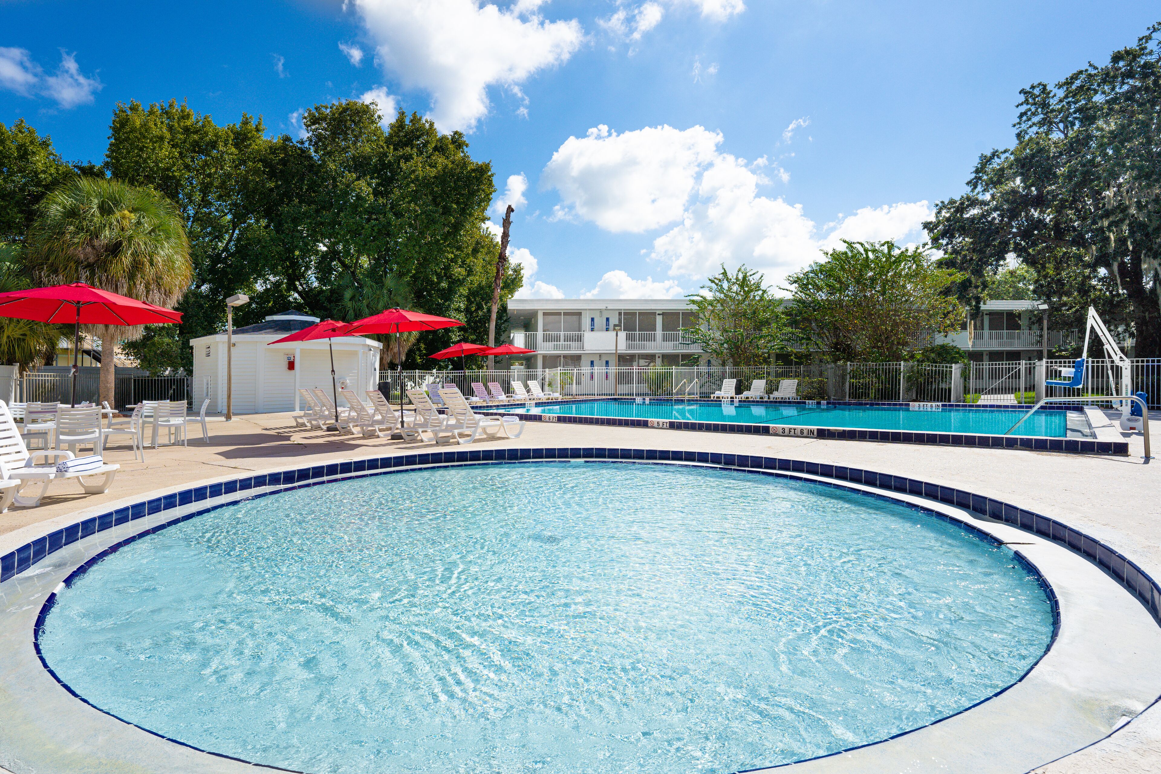 A swimming pool with lounge chairs and red umbrellas, surrounded by trees and a building with a balcony.