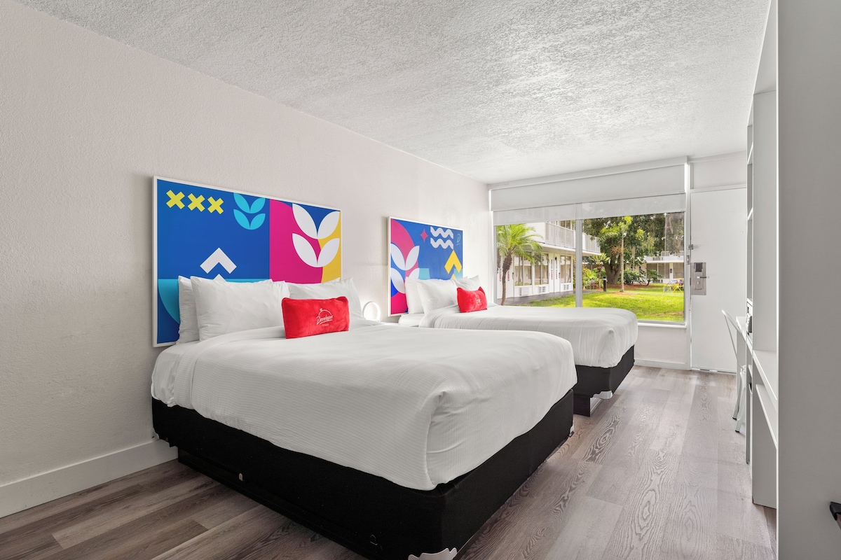 A hotel room with two beds, white bedding, red pillows, and colorful abstract wall art.