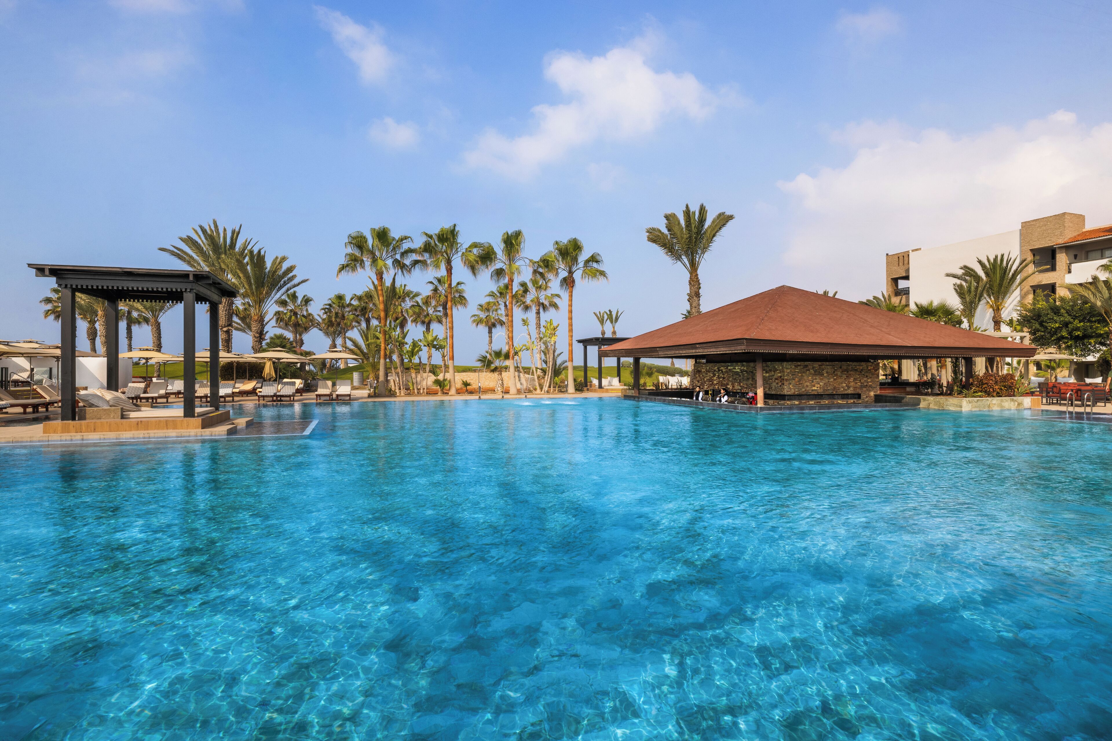 Ein klares, blaues Schwimmbecken mit Liegestühlen und Sonnenschirmen, umgeben von Palmen und einem Pavillon.