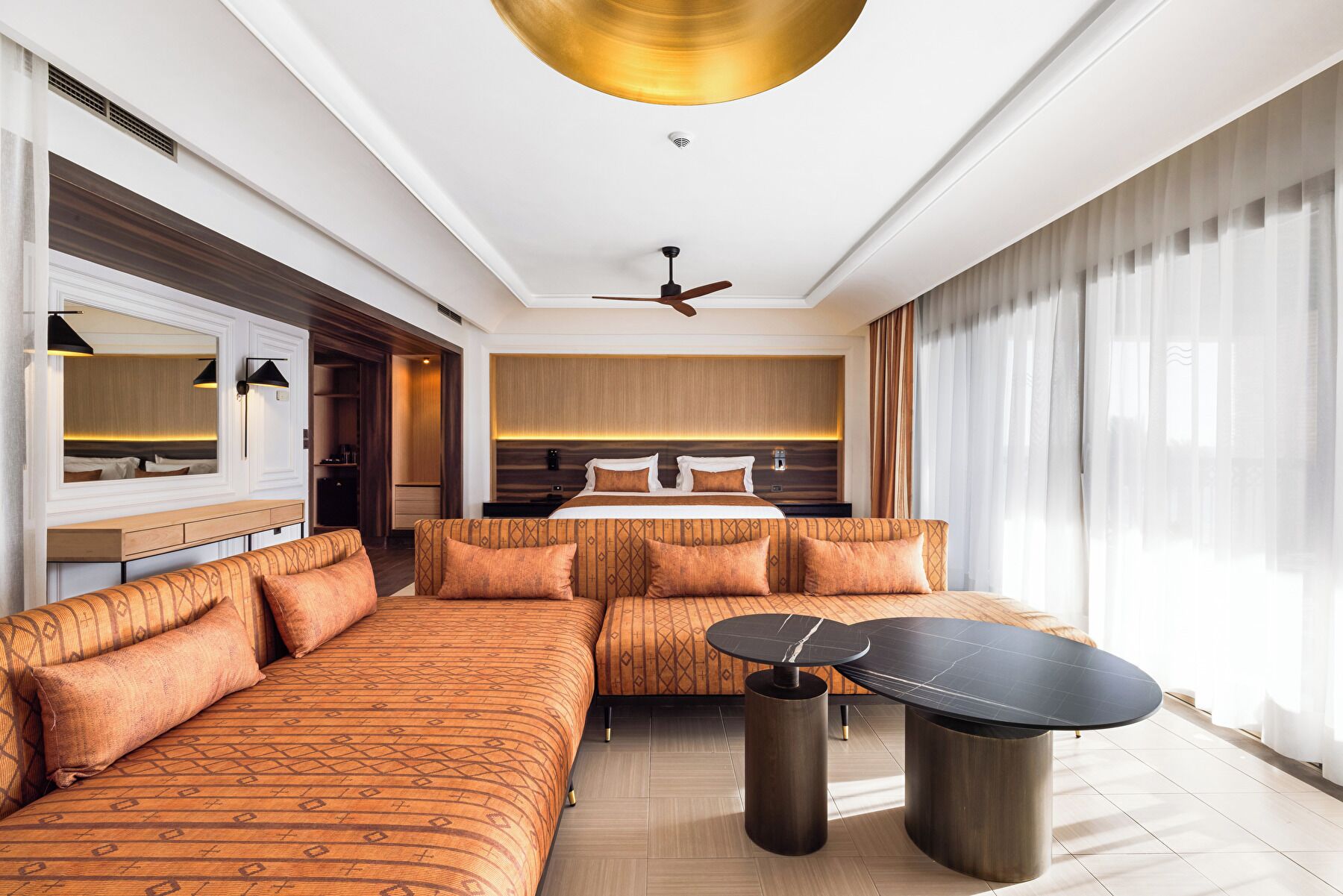 Una habitación de hotel moderna con un sofá grande, una mesa de comedor, una cama con cabecera, un ventilador de techo y ventanas amplias con cortinas.