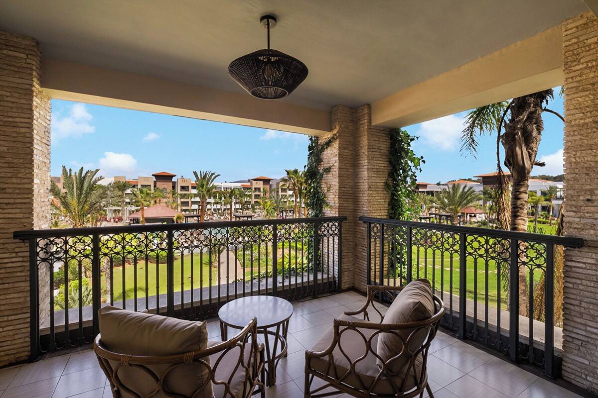 A balcony with wicker furniture, a round table, and a view of a resort with palm trees and buildings.