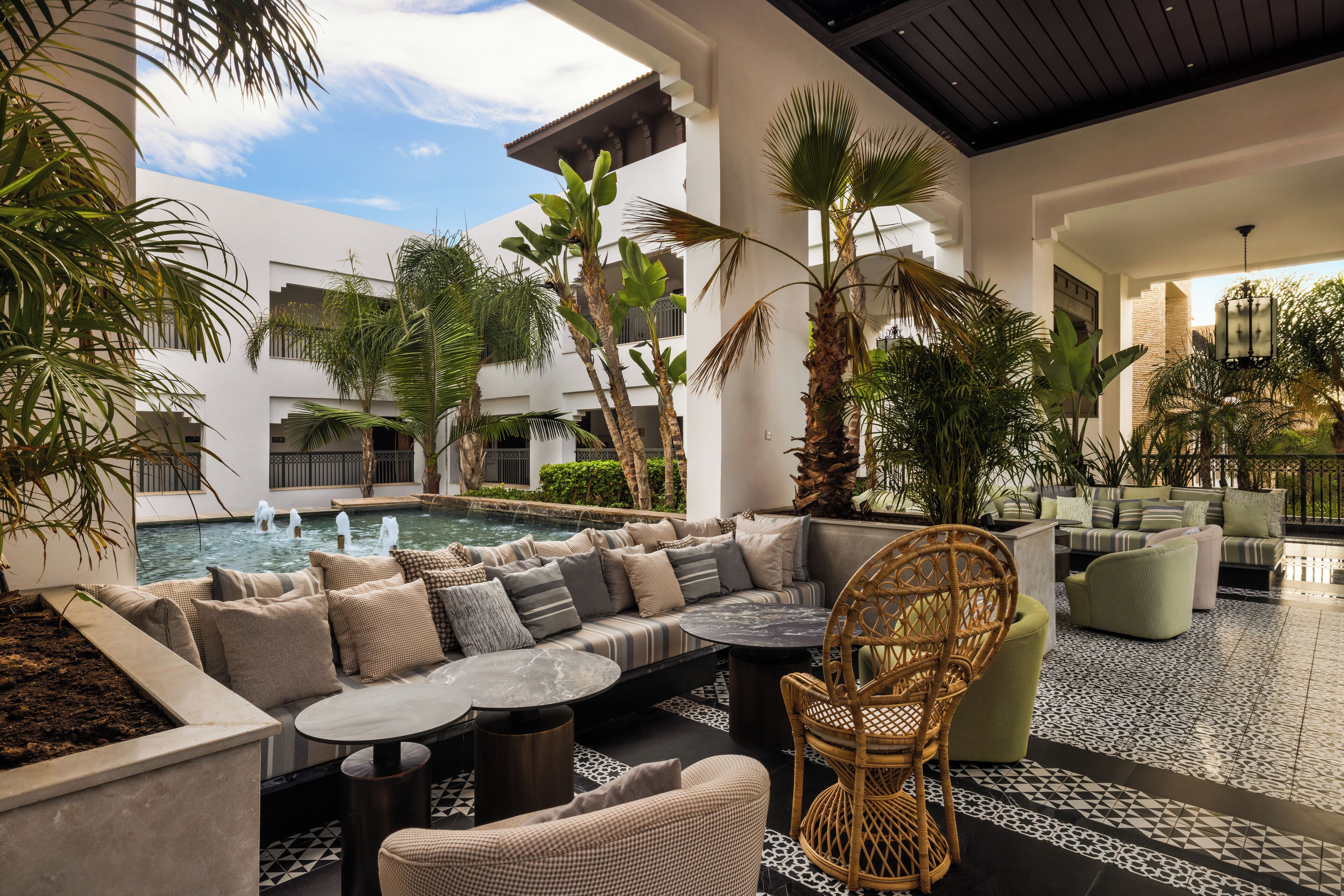 Zona de piscina con asientos, palmeras y vistas a un edificio con balcones.