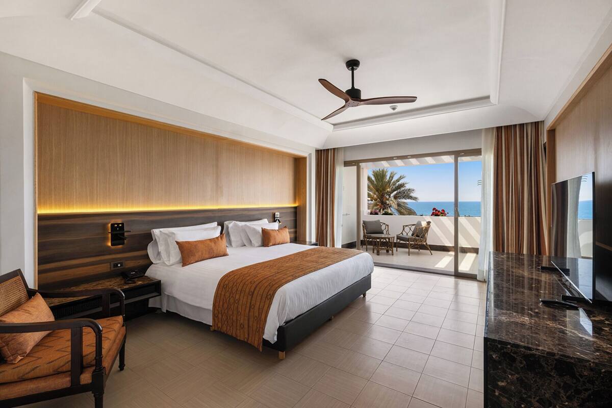 A hotel room with a bed, bedside table, chair, TV, and a balcony with a view of the sea.