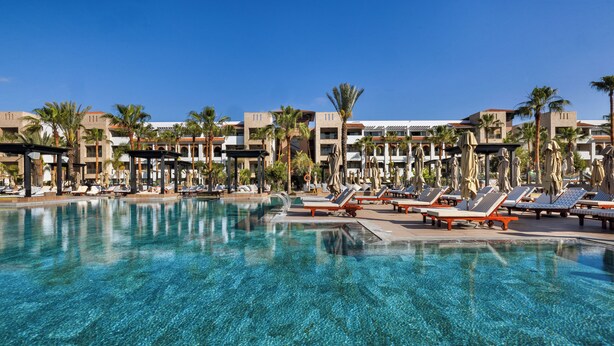 A resort pool with clear blue water, sun loungers, palm trees, and resort buildings in the background. - Hotel Riu Palace Tikida Agadir - All Inclusive (Agadir)
