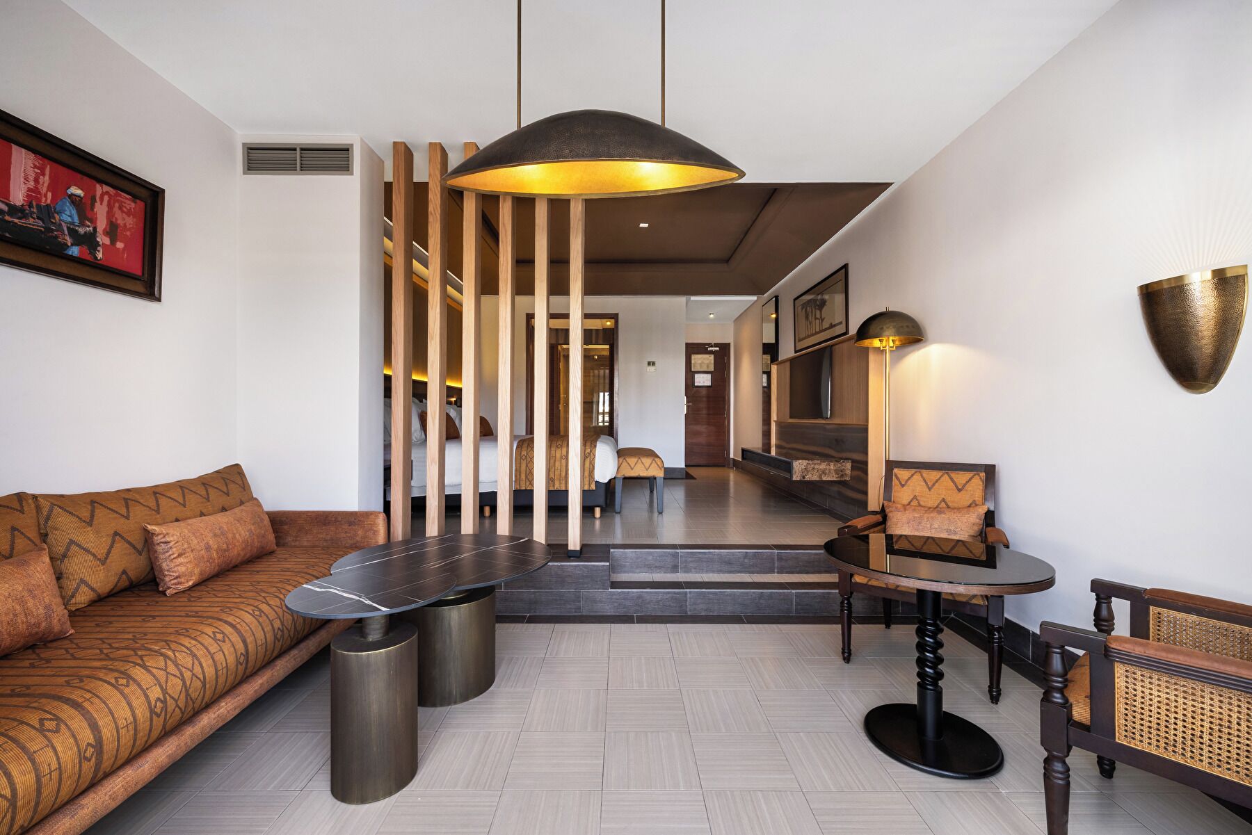 A modern hotel lobby with a sofa, a round table, and a chair. There is a wooden partition with vertical slats and a hanging pendant light.