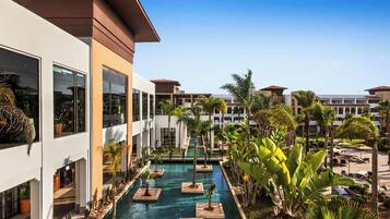 A modern hotel with a central swimming pool, surrounded by palm trees and lounge areas.