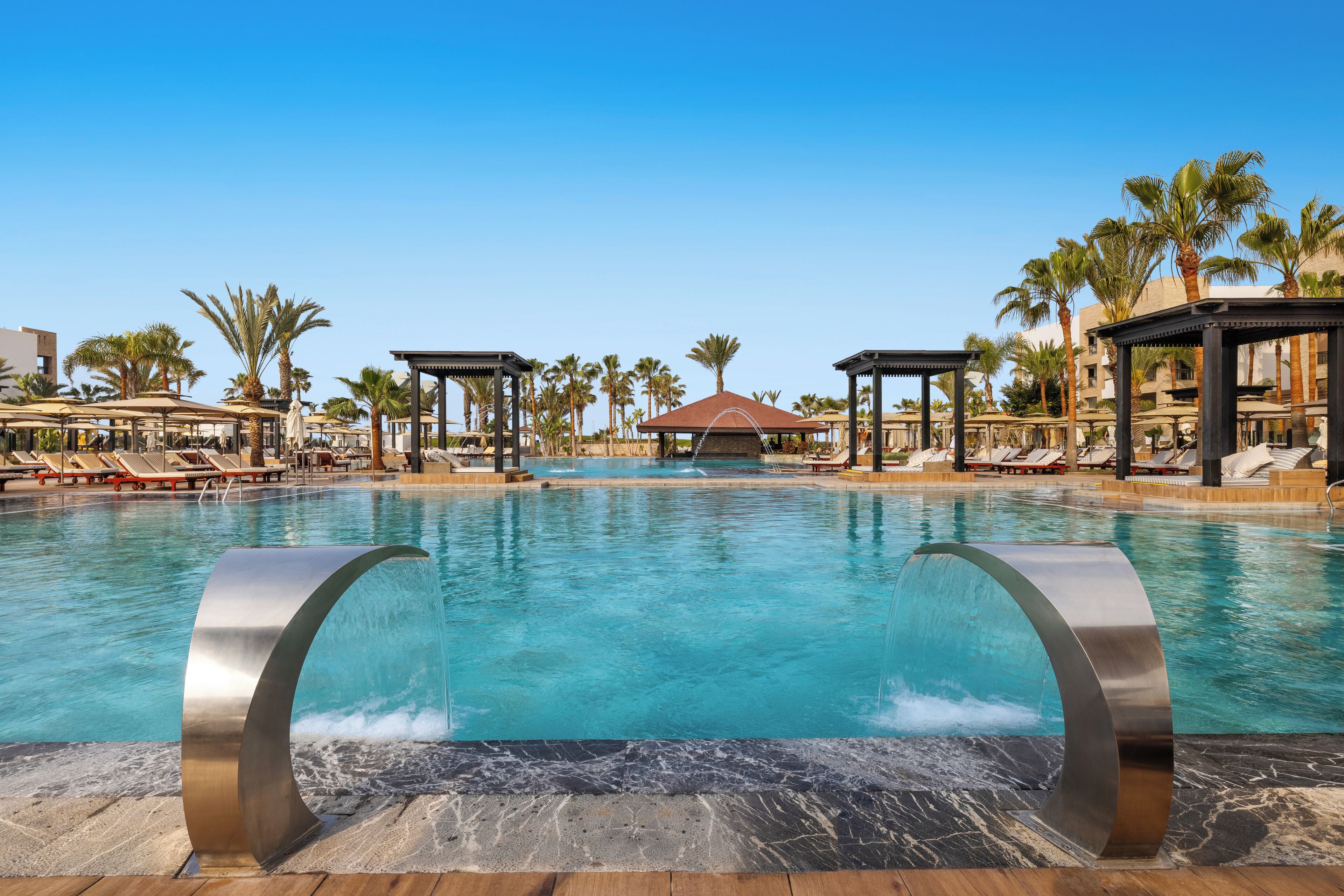 A swimming pool with two large metallic structures in the foreground, surrounded by lounge chairs and palm trees.
