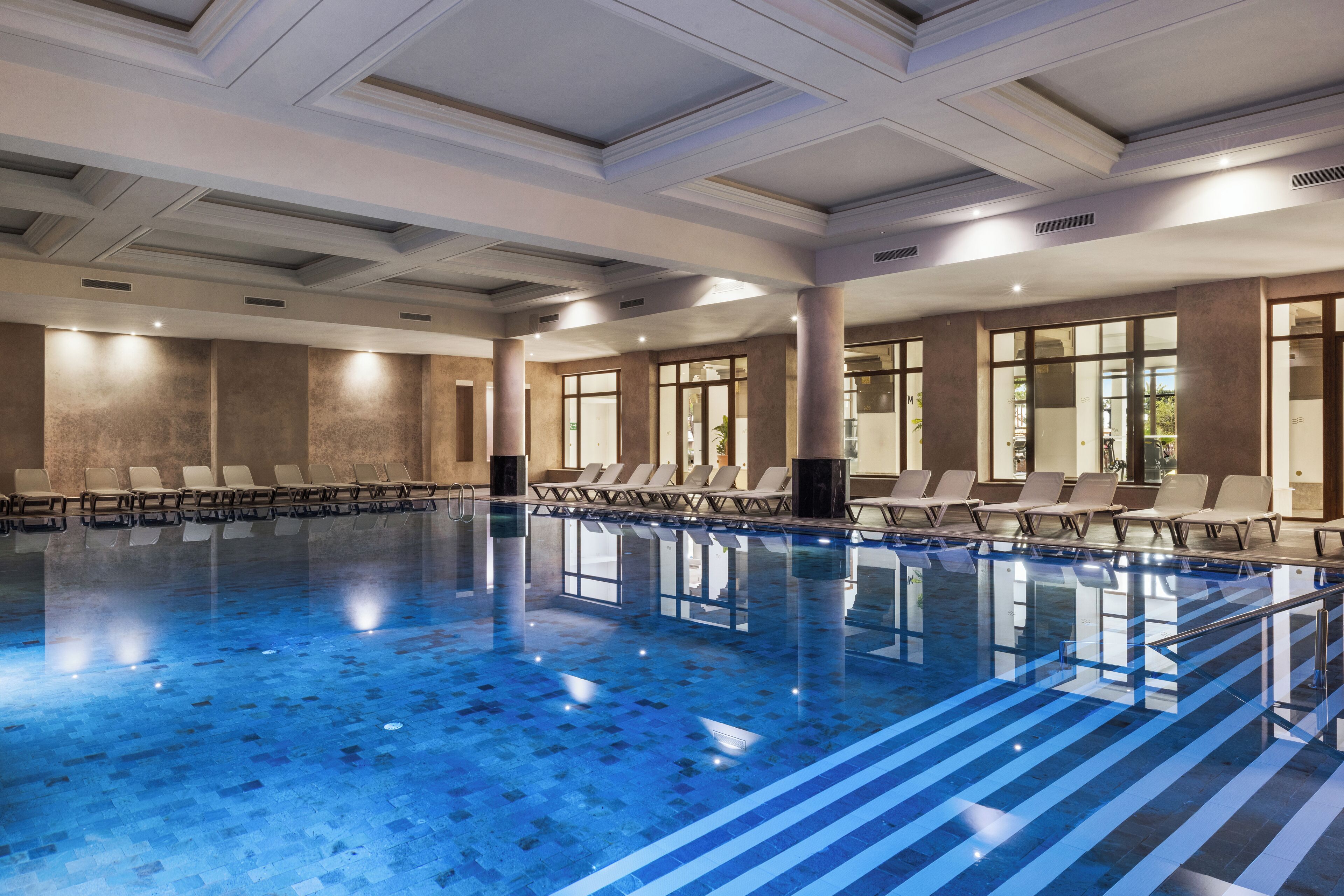 An indoor swimming pool with lounge chairs and a modern ceiling design.