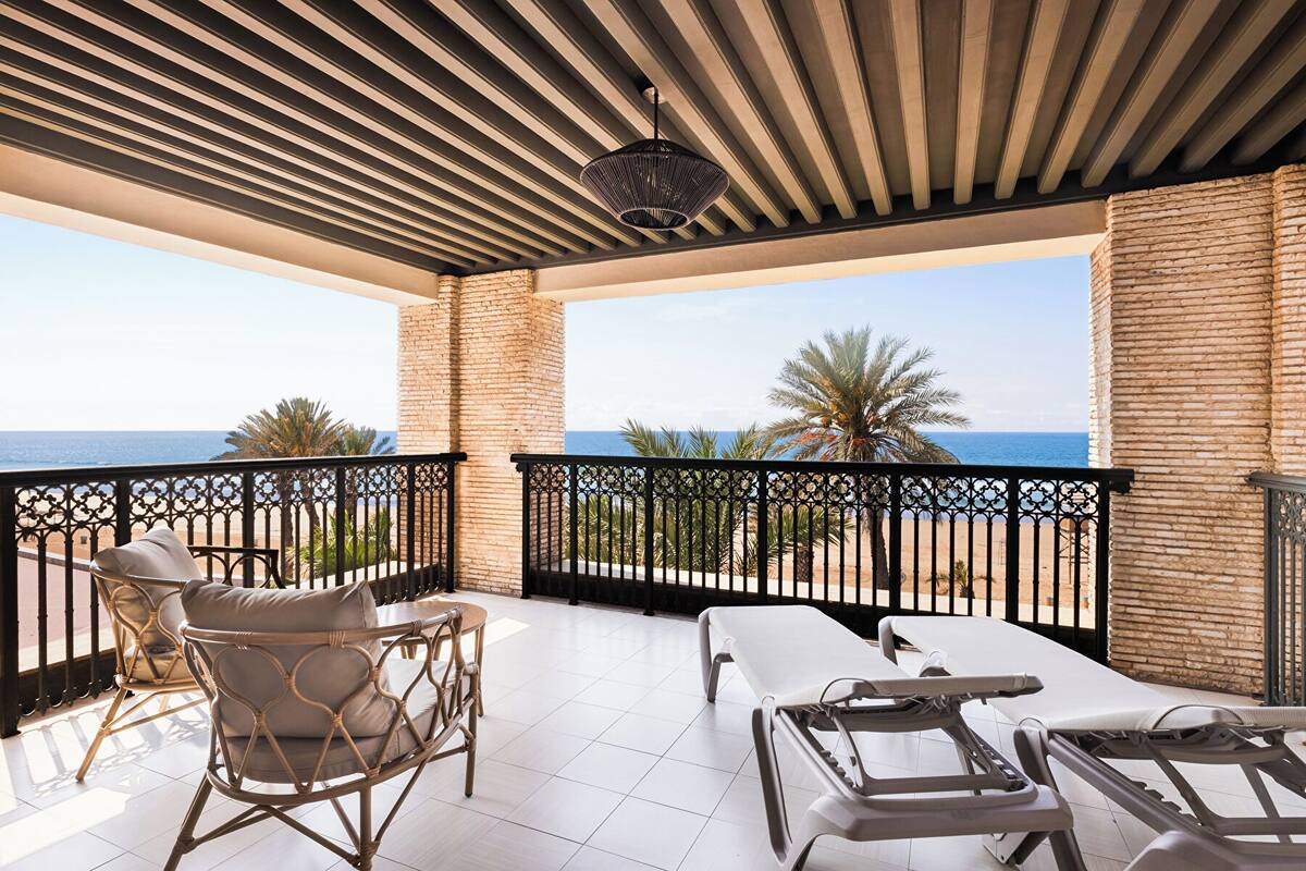A balcony with wicker chairs and a massage table, overlooking a beach and palm trees.