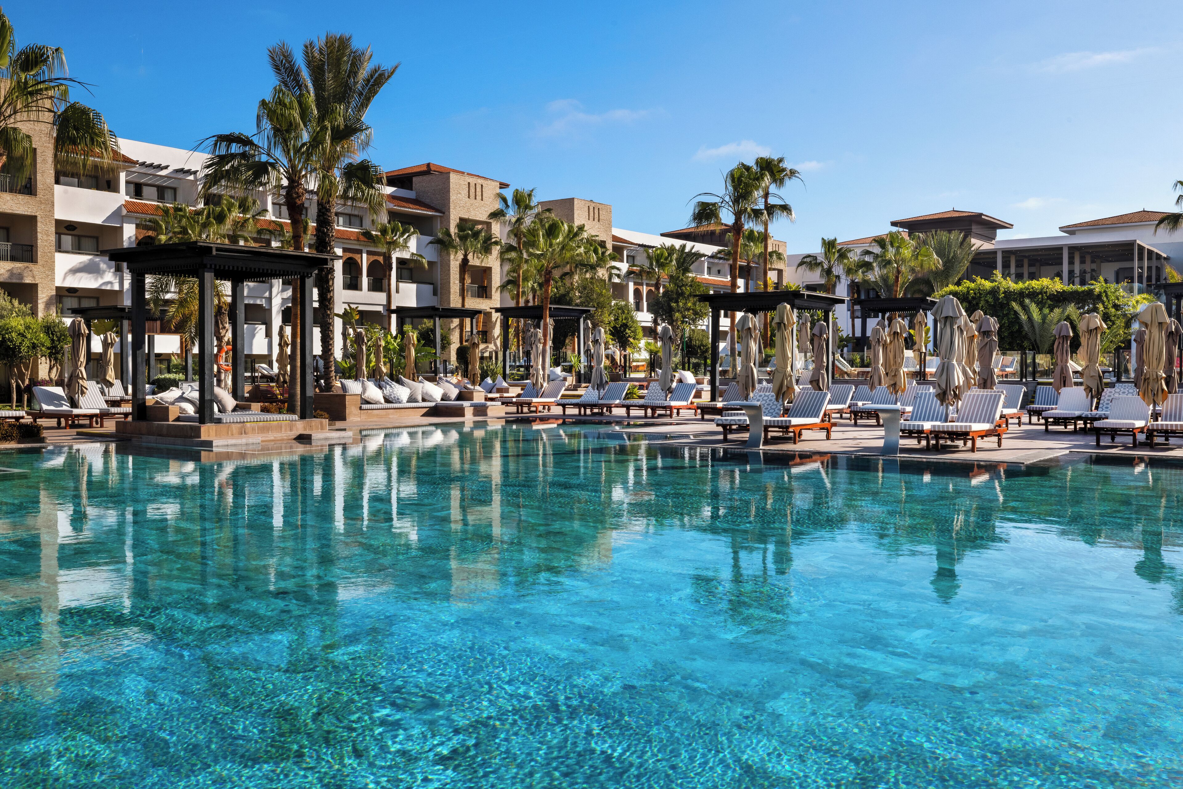 A clear pool with lounge chairs and umbrellas, surrounded by palm trees and hotel buildings.