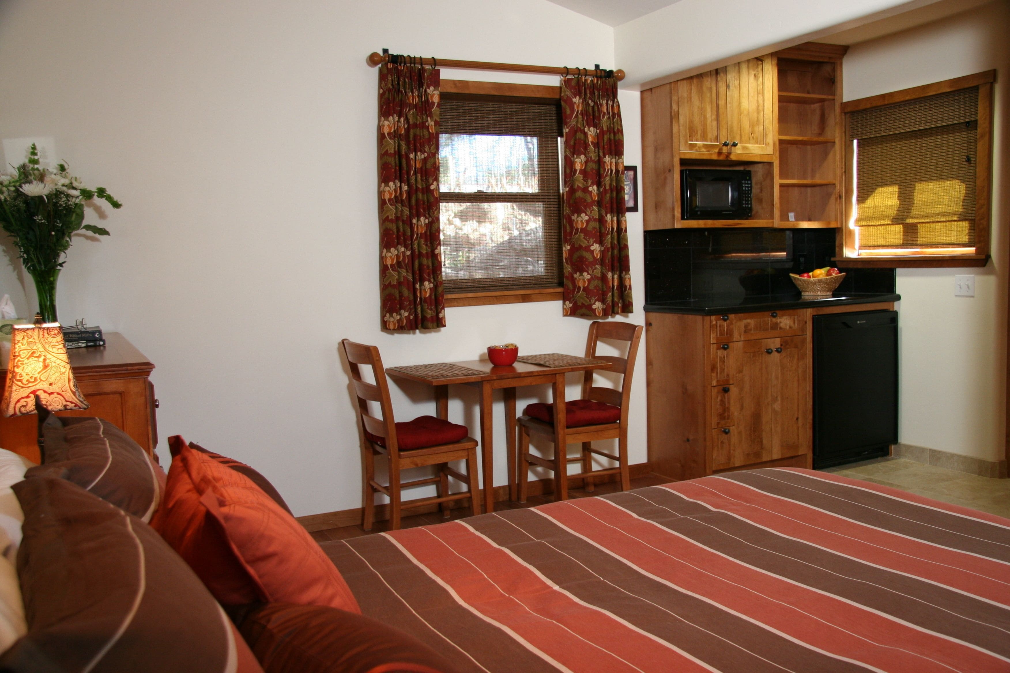 A bedroom with a bed, a bedside table with a lamp, a window with curtains, a small dining table with chairs, and a kitchenette with a microwave and sink.