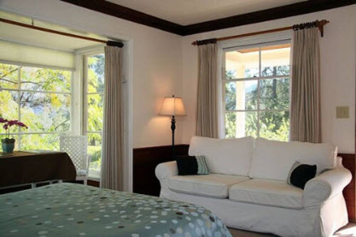 A room with a bed, a white sofa, a table with a lamp, and large windows with curtains.