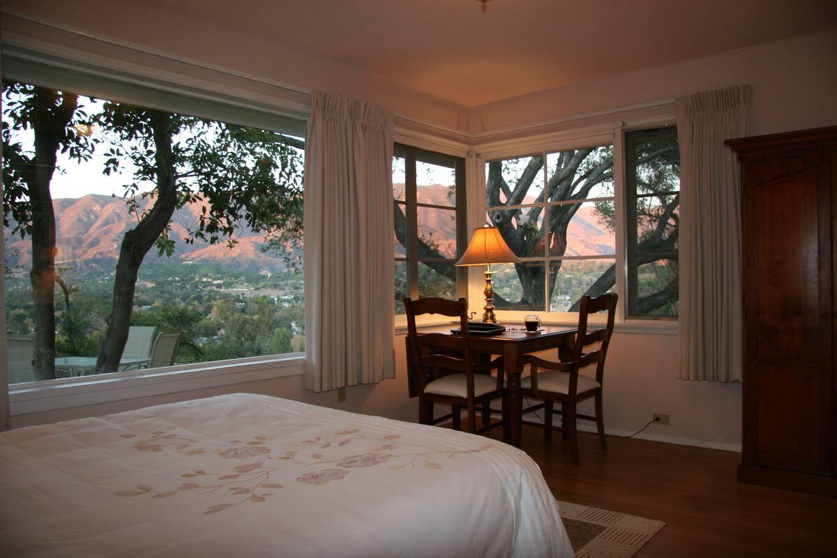 A bedroom with a large bed, a desk with chairs, and a view of mountains through the windows.