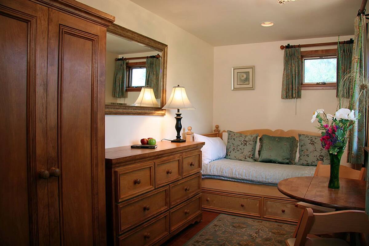 A bedroom with a wooden dresser, a bed with floral bedding, a bedside table with a lamp, and a window with curtains.