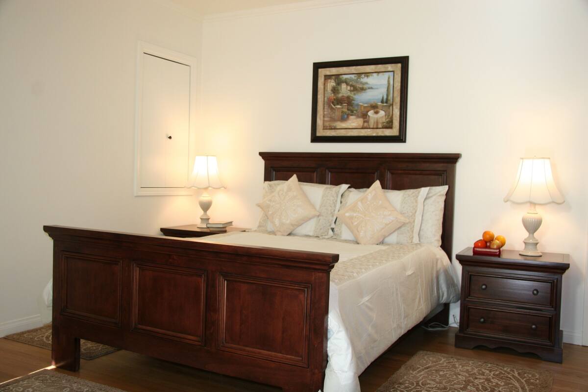 A bedroom with a wooden bed, two bedside tables with lamps, a framed picture on the wall, and a door.