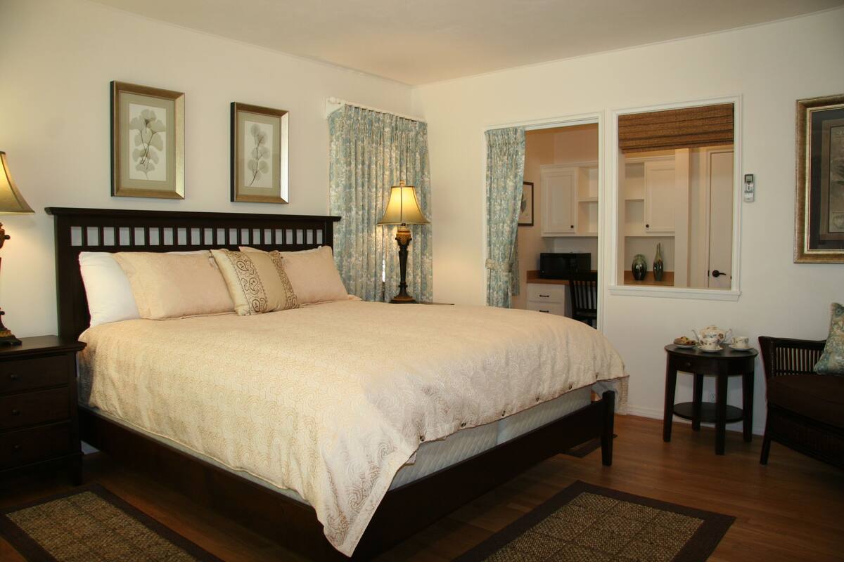 A bedroom with a large bed, a bedside table with a lamp, a small table with a teapot, and a chair.