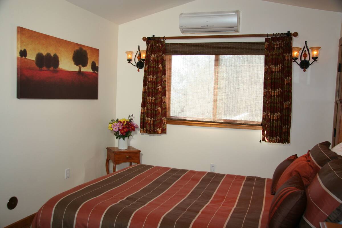 A bedroom with a bed, a window with curtains, a wall-mounted air conditioner, a nightstand with a vase of flowers, and a painting on the wall.