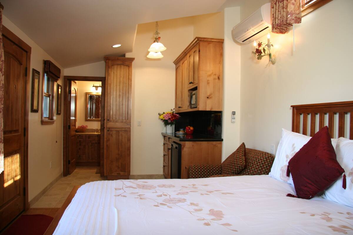 A bedroom with a large bed, wooden wardrobe, and a small kitchenette.