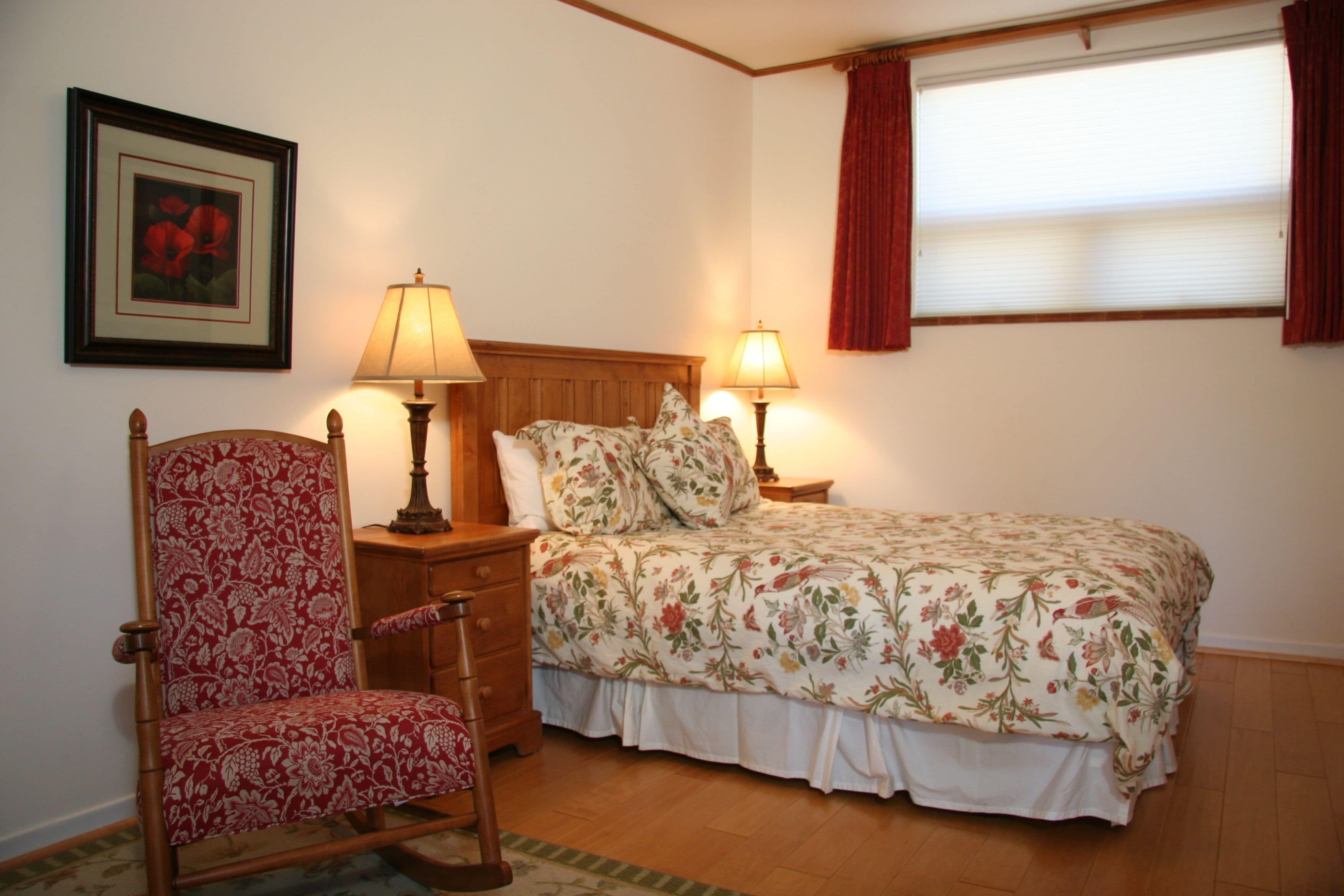 A bedroom with a wooden headboard, floral bedspread, two bedside lamps, a chair with a floral pattern, and a framed picture on the wall.