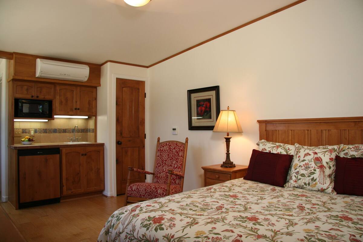 A bedroom with a bed, a chair, a bedside table, a lamp, and a kitchen area with a microwave and cabinets.