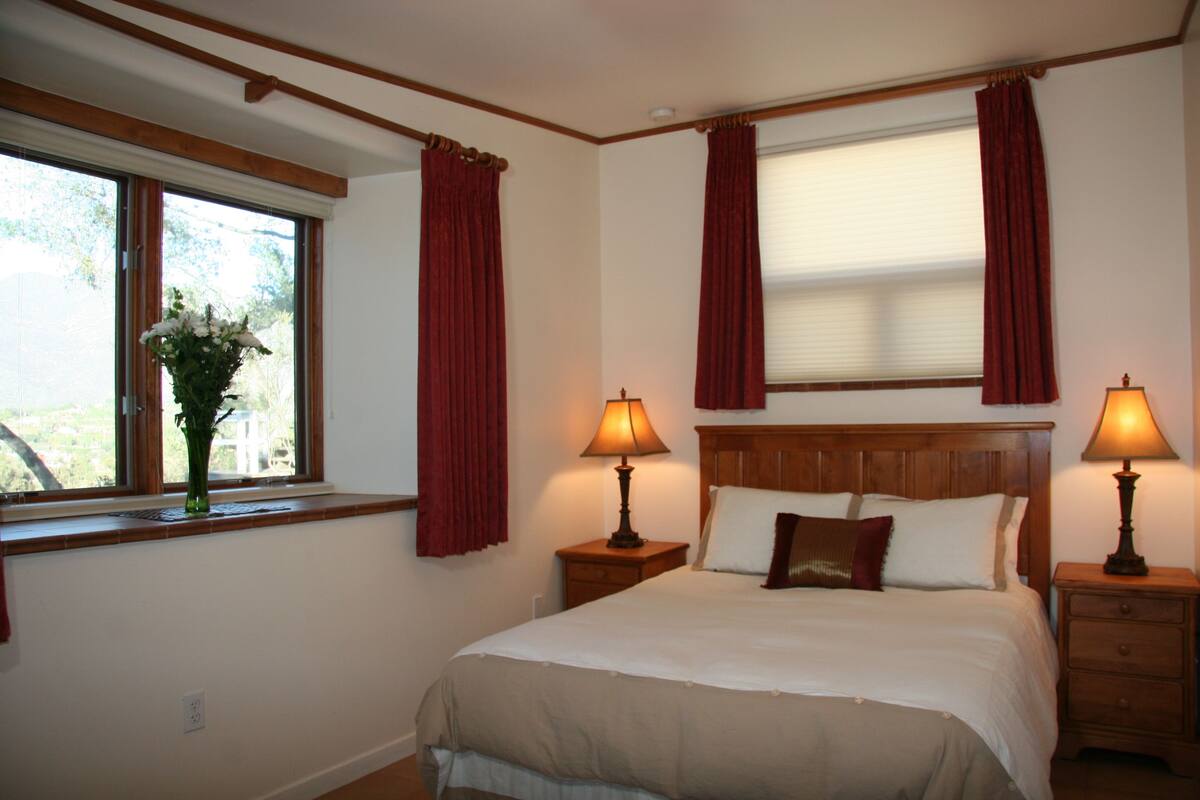 A bedroom with a large bed, two bedside tables with lamps, a window with curtains, and a vase with flowers on the windowsill.