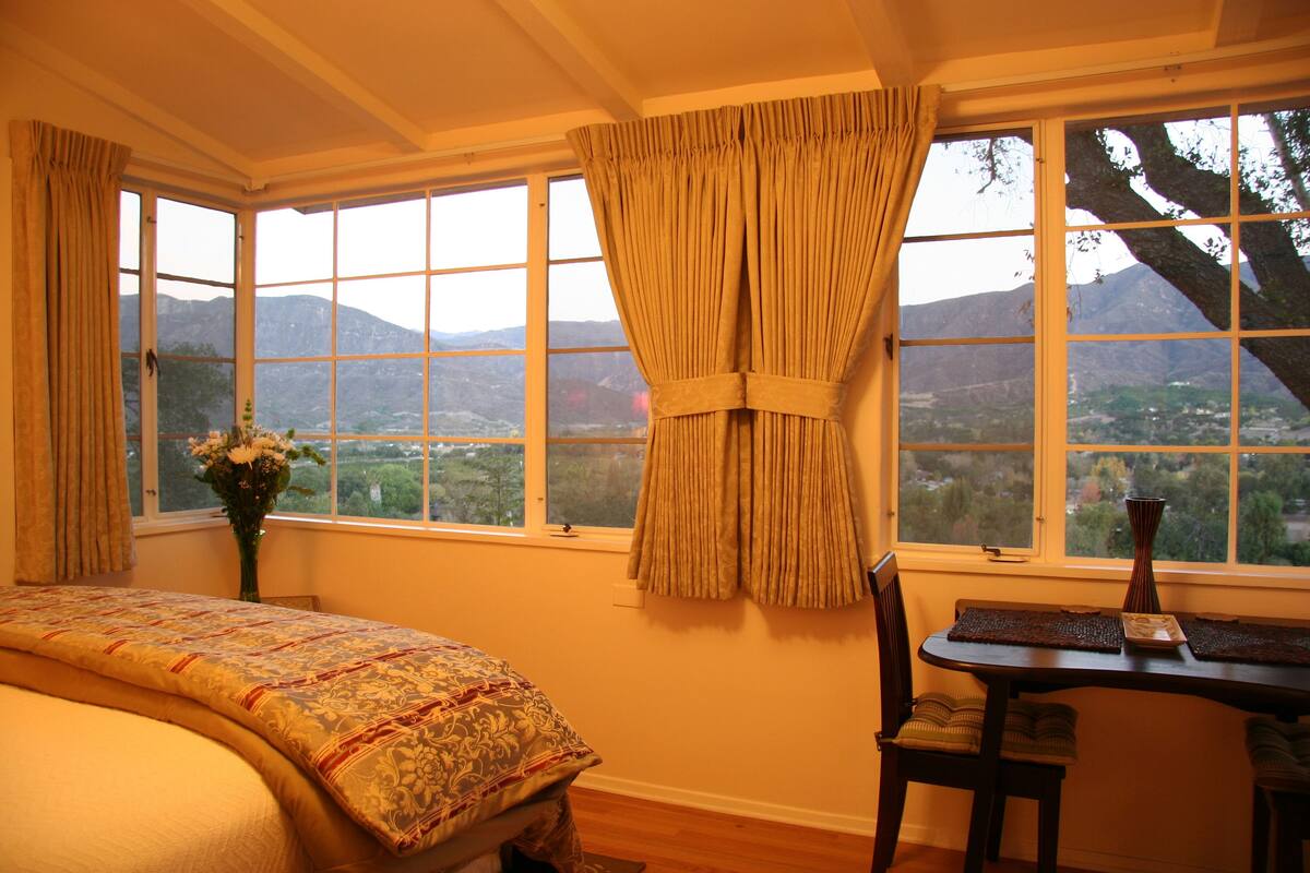 A bedroom with a large bed, a dining table with chairs, and a view of mountains through large windows.