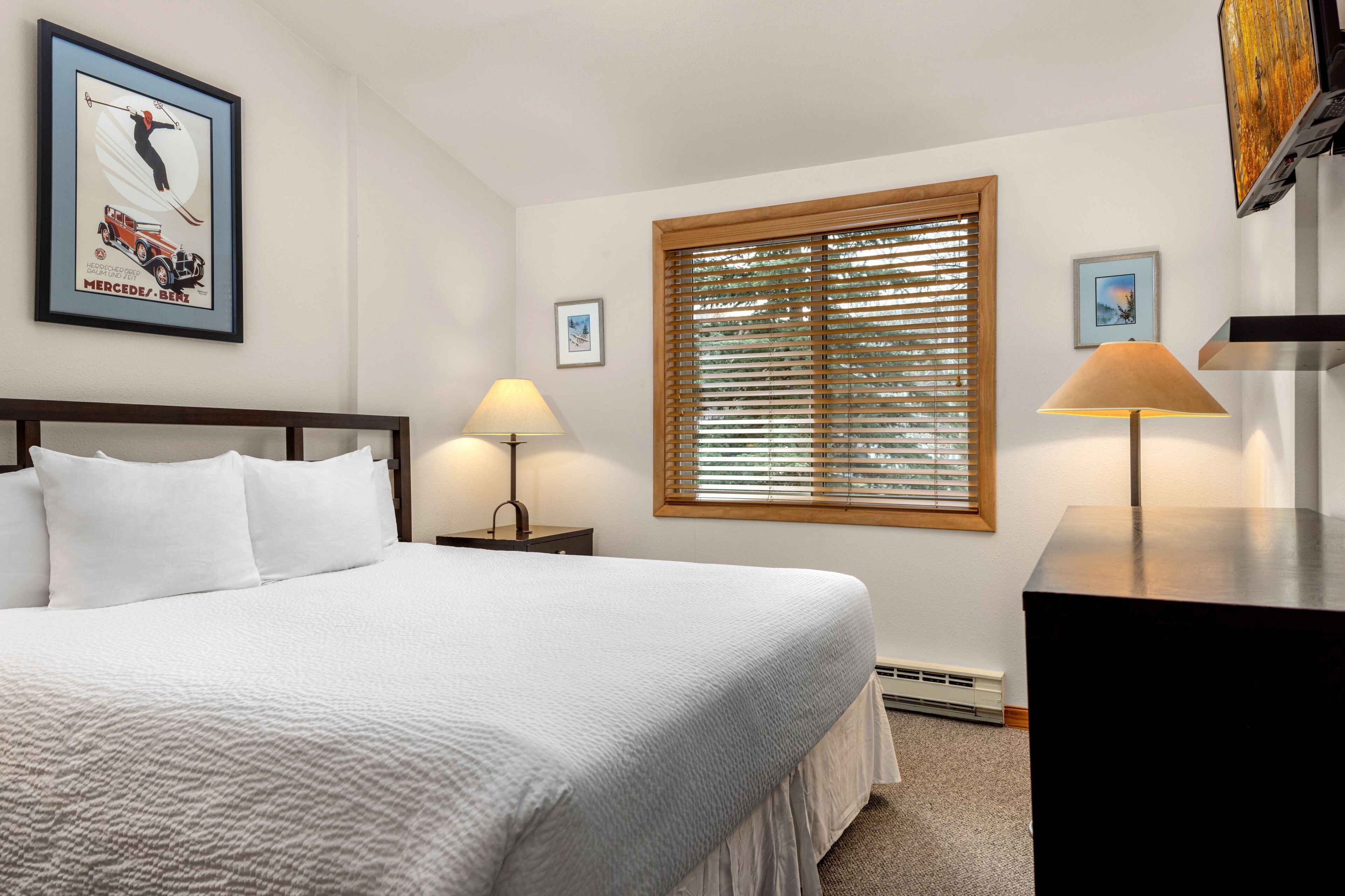 A neatly made bed with white linens, a wooden headboard, and two bedside lamps. A large window with blinds is on the wall, and a framed picture of a car is above the bed.