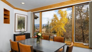 A dining area with a large table and six chairs, a vase with flowers, and a view of autumn trees through large windows. - Terracehouse - CoralTree Residence Collection (Snowmass Village)