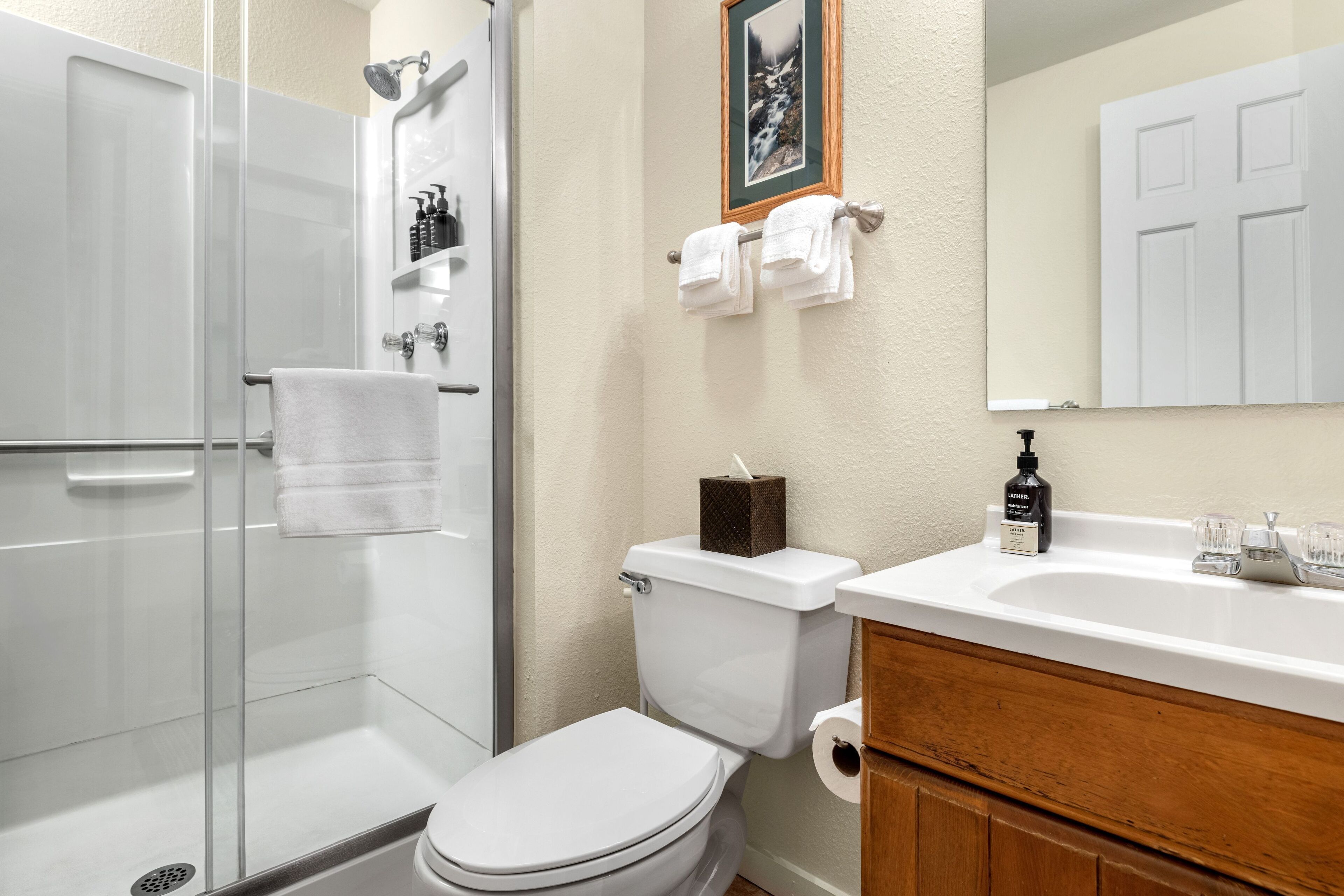 Een badkamer met een douchecabine van glas, een toilet, een wastafel met toiletartikelen en een lijst met een afbeelding aan de muur.