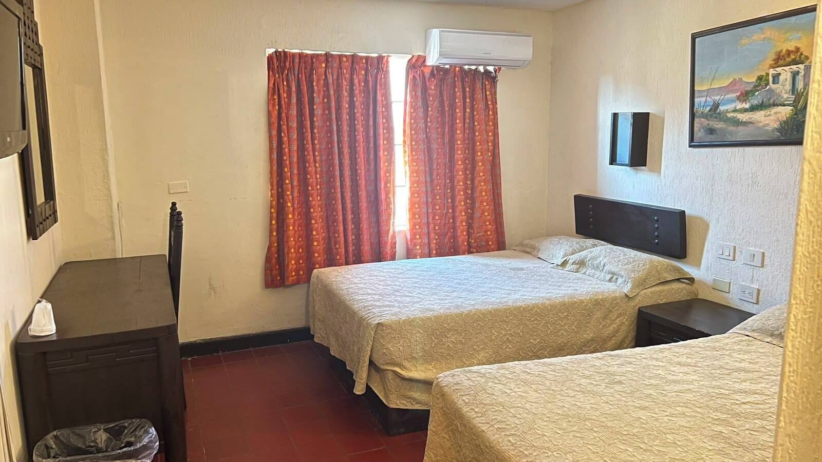 Ein Hotelzimmer mit zwei Betten, einem Schreibtisch, einem Mülleimer und einem Bild an der Wand.