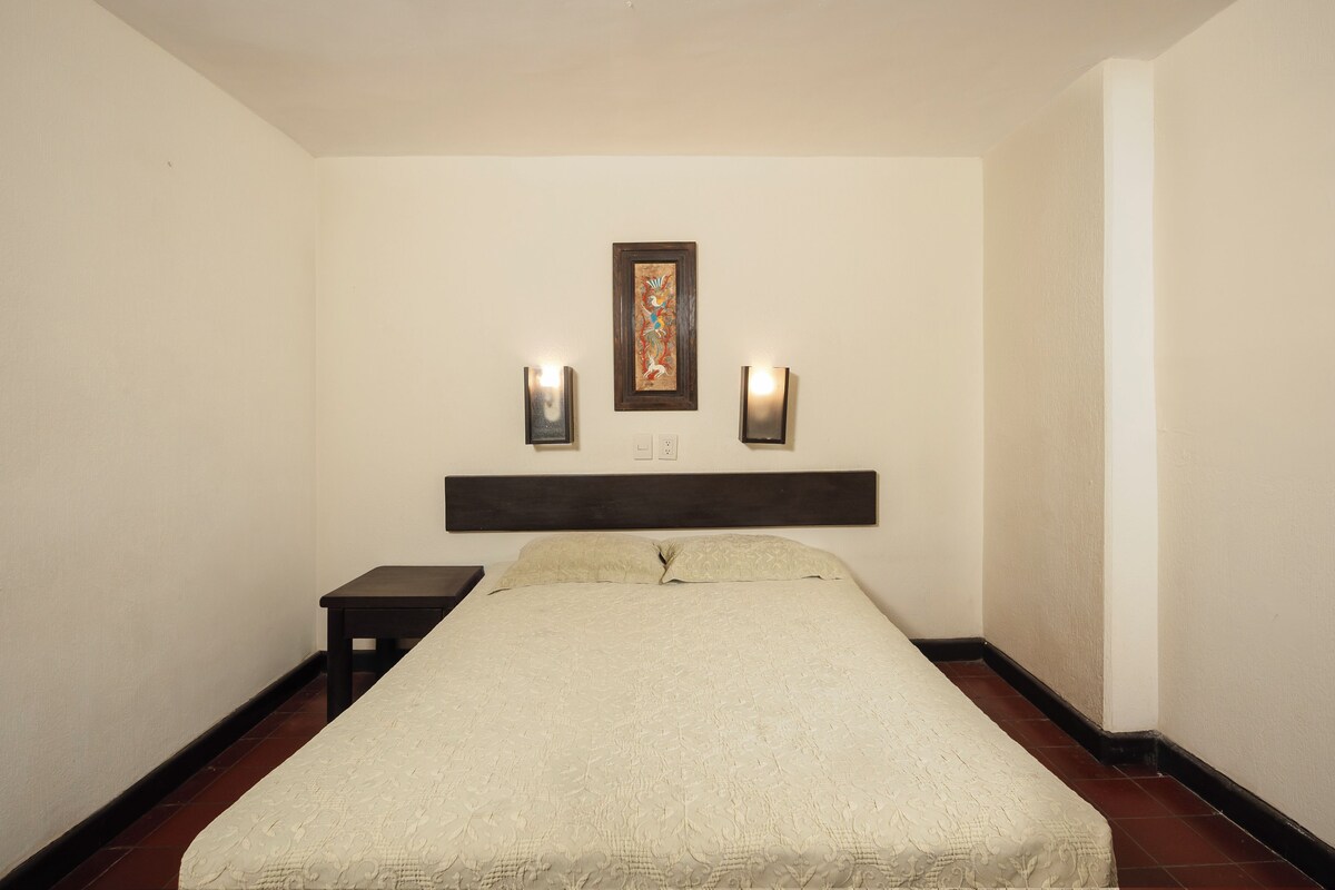 Habitación de hotel con una cama, una mesita de noche, dos lámparas de pared y un cuadro enmarcado sobre la cama.