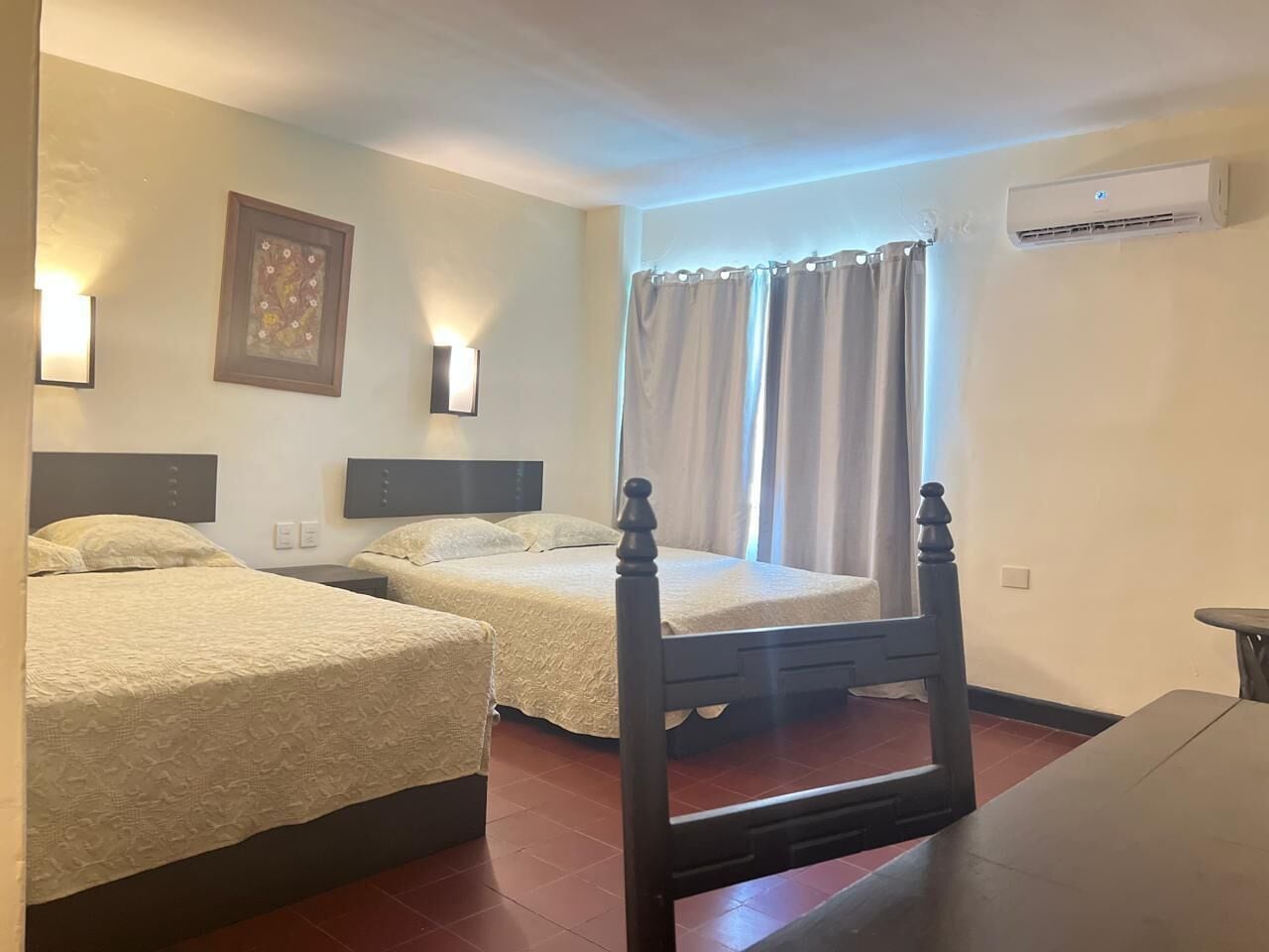 Habitación de hotel con dos camas, cabecero de madera, mesita de noche, aire acondicionado instalado en la pared y una ventana con cortinas.