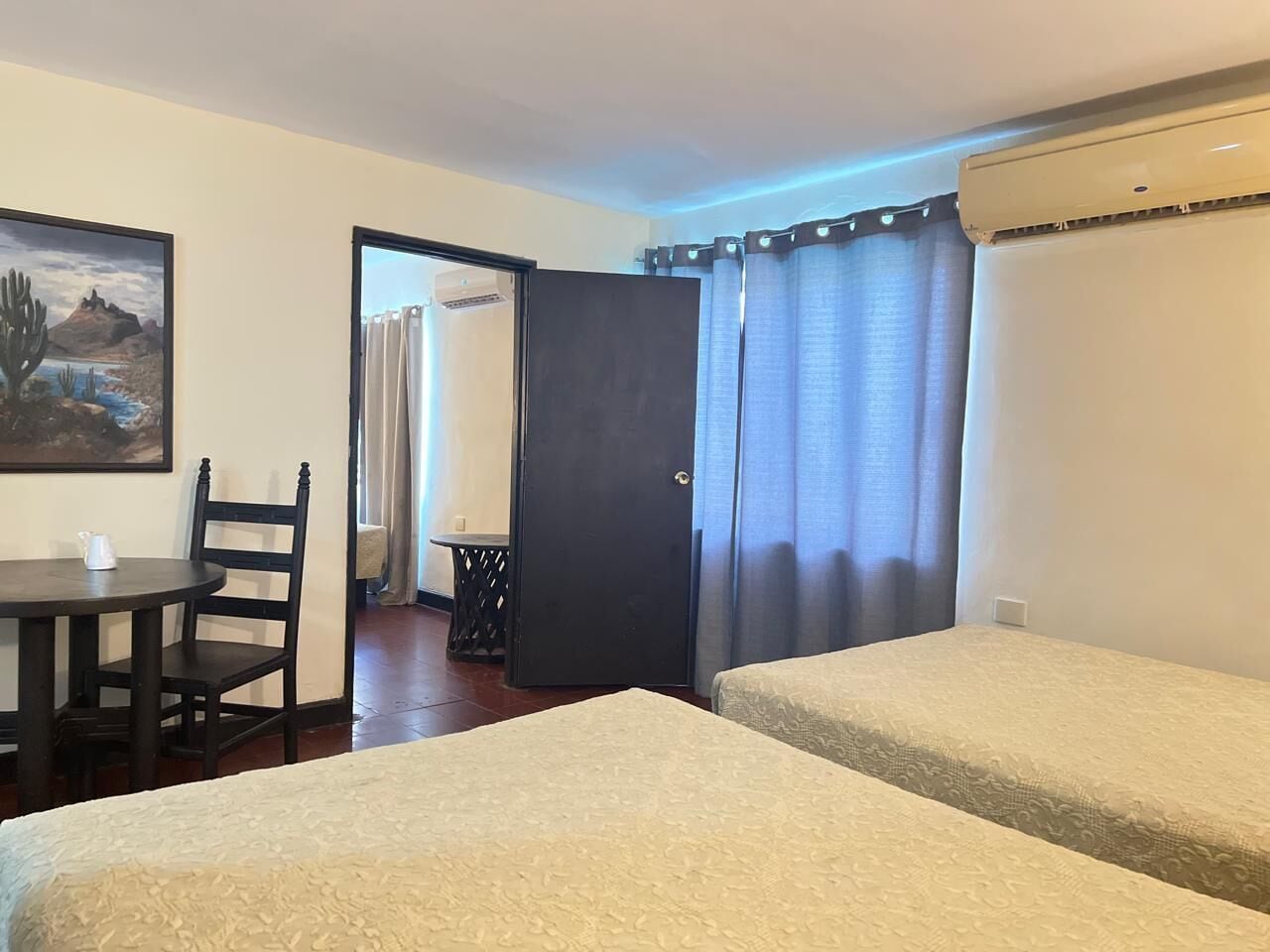 A hotel room with two single beds, a small table with a chair, a painting on the wall, and a door leading to another room.