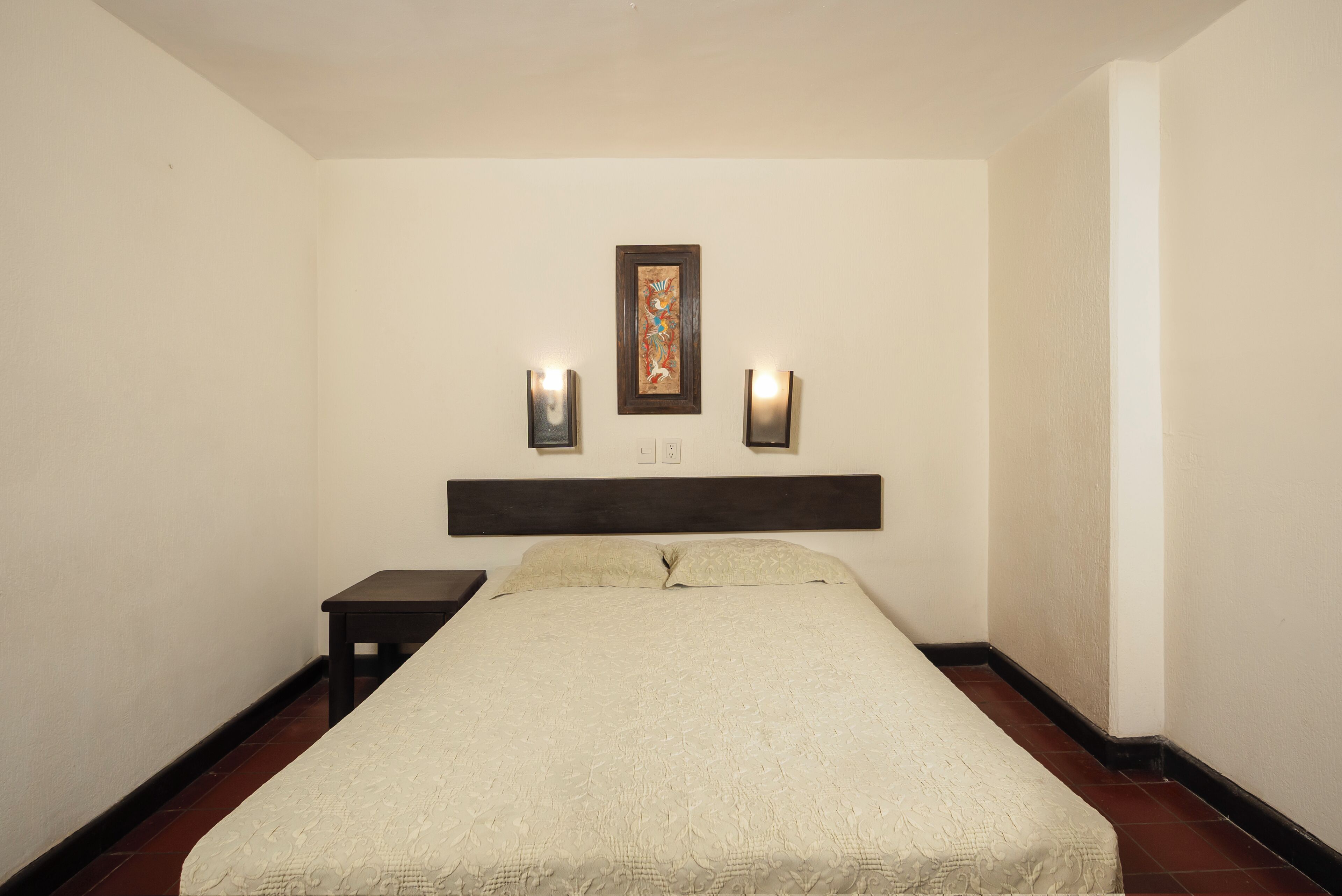 A hotel room with a bed, a nightstand, two wall-mounted lights, and a framed picture above the bed.