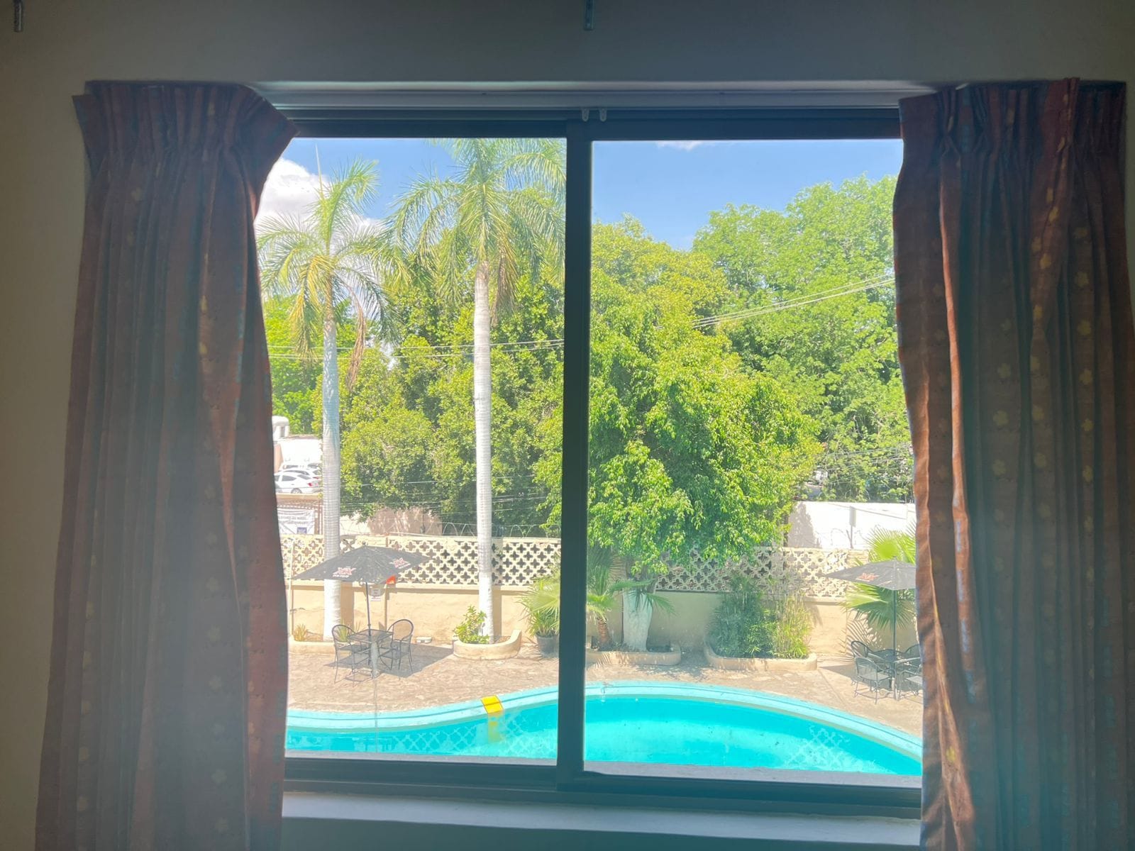 A view through a window with curtains, featuring a swimming pool, outdoor furniture, and lush greenery.