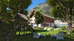 A rustic wooden playhouse with a slide, surrounded by a grassy area with white lounge chairs and a tree. - Hotel Les Touristes (Abondance)