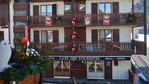 A building with a sign reading "CAFE DES TOURISTES" and "STELLA ARTOIS" on the ground floor. There are balconies with decorations and a large snowman figure on the left. - Hotel Les Touristes (Abondance)