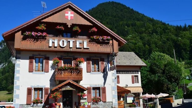 A hotel with a prominent sign, a balcony with flowers, and a mountain backdrop. - Hotel Les Touristes (Abondance)
